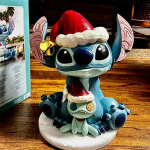 May include: A blue and white figurine of a cartoon character wearing a red Santa hat and a gold bell. The figurine is on a white base and has a smaller matching figurine. The figurine is a Christmas decoration.