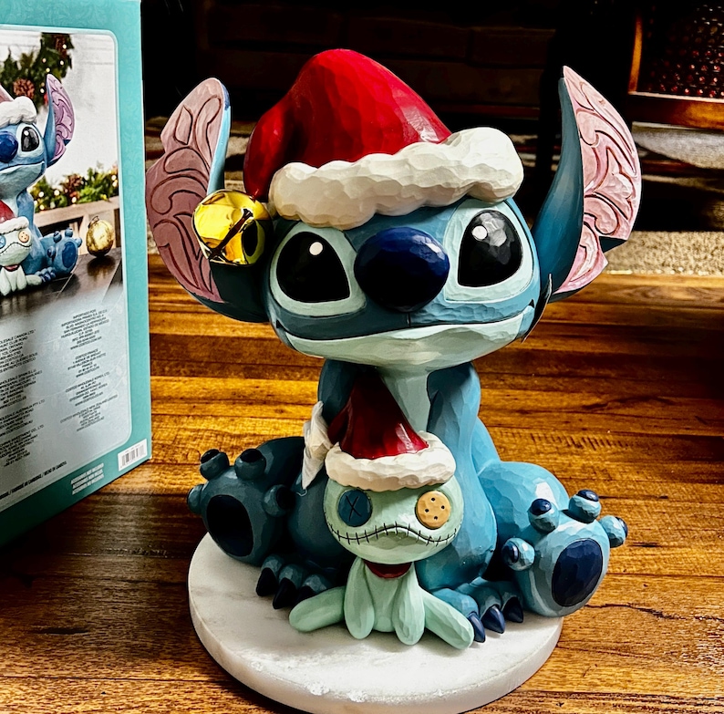 May include: A blue and white figurine of a cartoon character wearing a red Santa hat and a gold bell. The figurine is on a white base and has a smaller matching figurine. The figurine is a Christmas decoration.