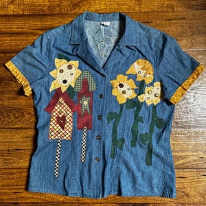 May include: Blue denim short-sleeve shirt with decorative appliqués. The shirt features appliquéd sunflowers, birdhouses, and stems. The cuffs have a yellow and gold patterned trim. The shirt has a button-down front and a collared neckline.