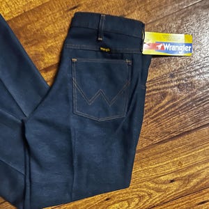 Może przedstawiać: Ciemnoniebieskie dżinsy z dołączoną metką Wrangler. Dżinsy mają klasyczny pięciokieszeniowy design z charakterystycznym szwem w kształcie litery W na tylnych kieszeniach. Dżinsy są złożone na drewnianej powierzchni.