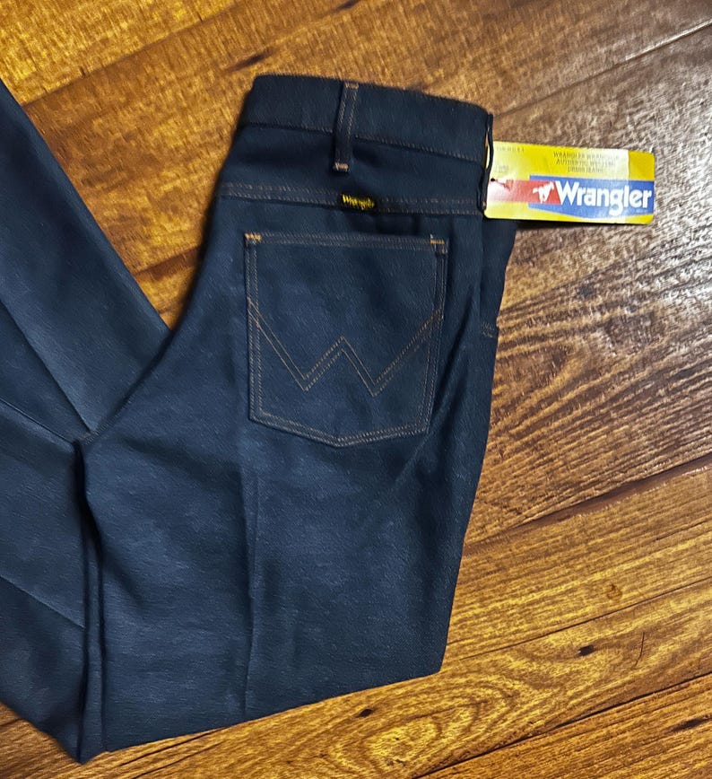 Może przedstawiać: Ciemnoniebieskie dżinsy z dołączoną metką Wrangler. Dżinsy mają klasyczny pięciokieszeniowy design z charakterystycznym szwem w kształcie litery W na tylnych kieszeniach. Dżinsy są złożone na drewnianej powierzchni.