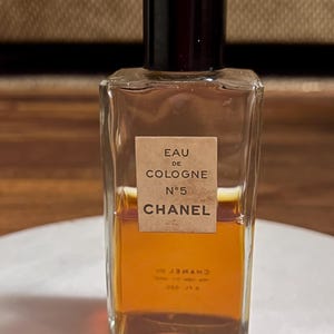 May include: A rectangular glass bottle of Chanel No. 5 Eau de Cologne. The bottle has a black cap and a label with the product name. The liquid inside is amber-colored. The bottle is on a white surface.
