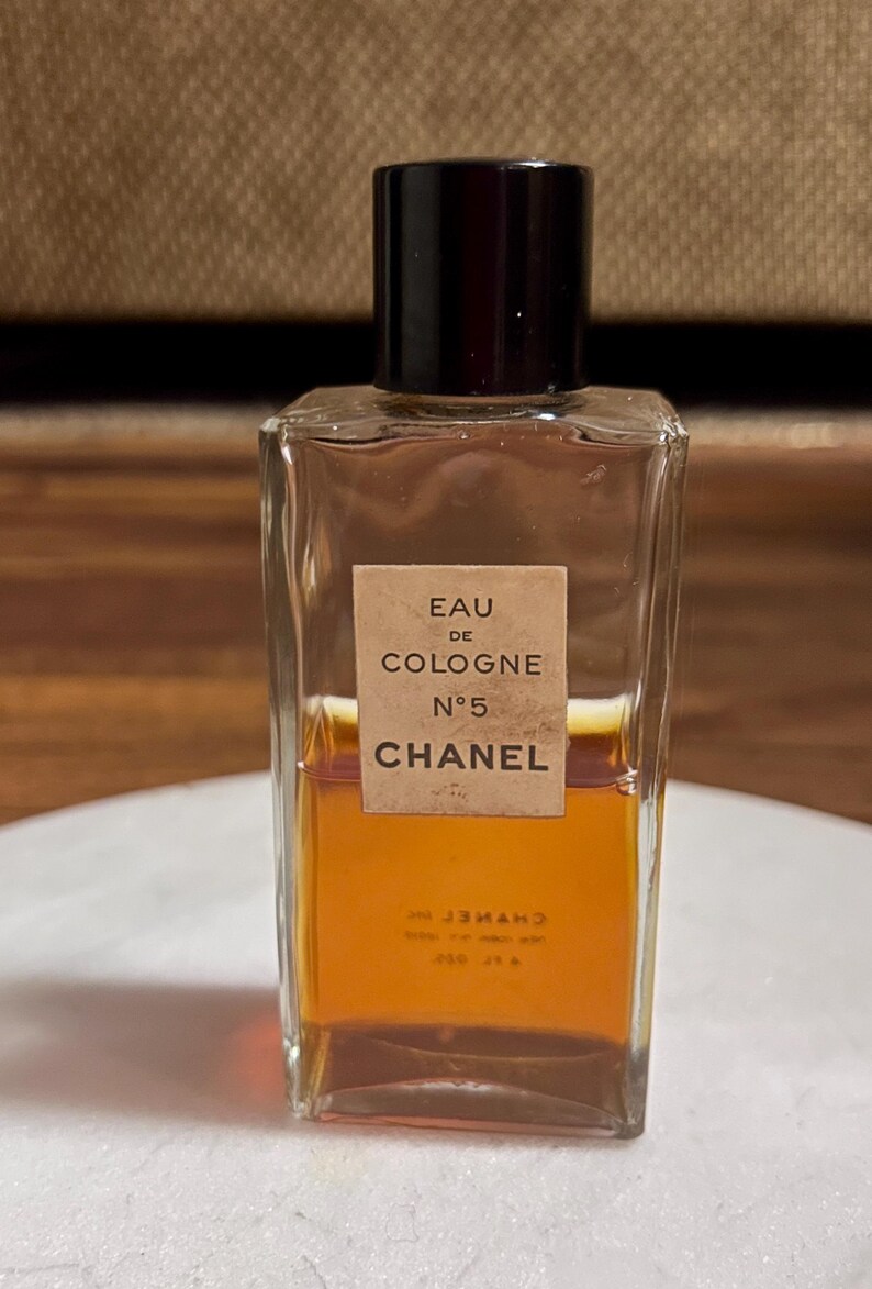 May include: A rectangular glass bottle of Chanel No. 5 Eau de Cologne. The bottle has a black cap and a label with the product name. The liquid inside is amber-colored. The bottle is on a white surface.