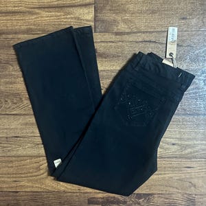 May include: Black denim jeans with a classic five-pocket design. The jeans feature a decorative pattern on the back pocket and a tag attached to the waistband. The pants are laid flat on a wooden surface.