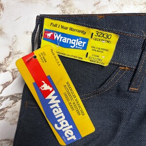 Może przedstawiać: Zbliżenie ciemnoniebieskich dżins&oacute;w Wrangler z dołączonymi ż&oacute;łtymi metkami. Metki wyświetlają logo Wrangler, rozmiar 32x30 (81x75cm) i inne szczeg&oacute;ły. Dżinsy są wyprodukowane w USA i składają się w 80% z poliestru i 20% z rayonu.