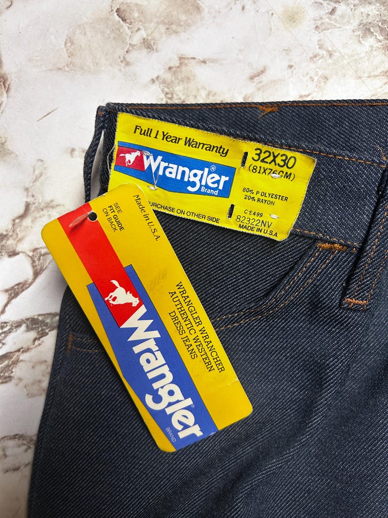 Może przedstawiać: Zbliżenie ciemnoniebieskich dżins&oacute;w Wrangler z dołączonymi ż&oacute;łtymi metkami. Metki wyświetlają logo Wrangler, rozmiar 32x30 (81x75cm) i inne szczeg&oacute;ły. Dżinsy są wyprodukowane w USA i składają się w 80% z poliestru i 20% z rayonu.