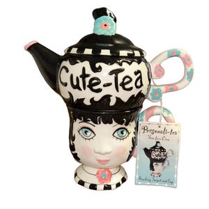 May include: A whimsical ceramic teapot and cup set. The teapot is black and white with the words "Cute-Tea" and a floral design. The cup is designed as a face with blue eyes and a pink flower. A tag reads "Personali-tea Tea for One".