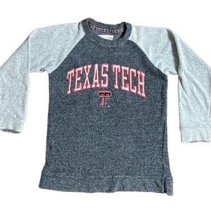 May include: A gray and charcoal raglan-sleeve sweatshirt with the words "TEXAS TECH" in red and white lettering across the chest. The letter "T" is below the words. The sleeves are light gray.