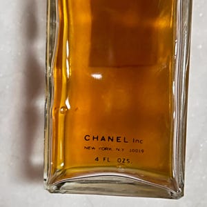 May include: A rectangular glass bottle filled with a golden-brown liquid. The bottom of the bottle features the text "CHANEL Inc NEW YORK, NY 10019" and "4 FL OZS."