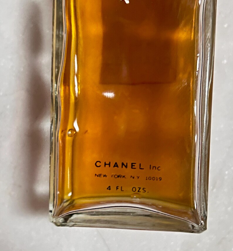 May include: A rectangular glass bottle filled with a golden-brown liquid. The bottom of the bottle features the text "CHANEL Inc NEW YORK, NY 10019" and "4 FL OZS."