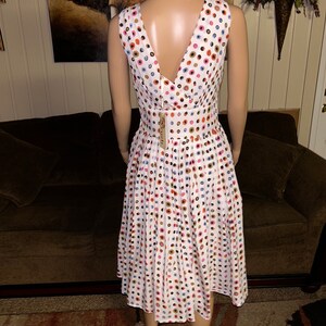 May include: White sleeveless dress with a V-neck back, adorned with a pattern of colorful polka dots. The dress features a pleated skirt and a fitted waist. A tag is attached to the back of the dress.