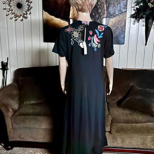 Puede incluir: Un vestido largo negro de manga corta con bordados coloridos de flores y fresas en la parte superior de la espalda. La prenda tiene cuello redondo, una etiqueta sujeta al cuello y un dobladillo con volantes, visto desde atr&aacute;s en un maniqu&iacute;.