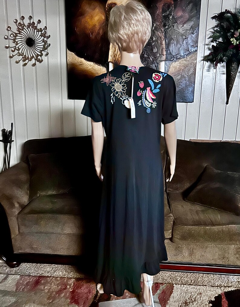 Puede incluir: Un vestido largo negro de manga corta con bordados coloridos de flores y fresas en la parte superior de la espalda. La prenda tiene cuello redondo, una etiqueta sujeta al cuello y un dobladillo con volantes, visto desde atr&aacute;s en un maniqu&iacute;.