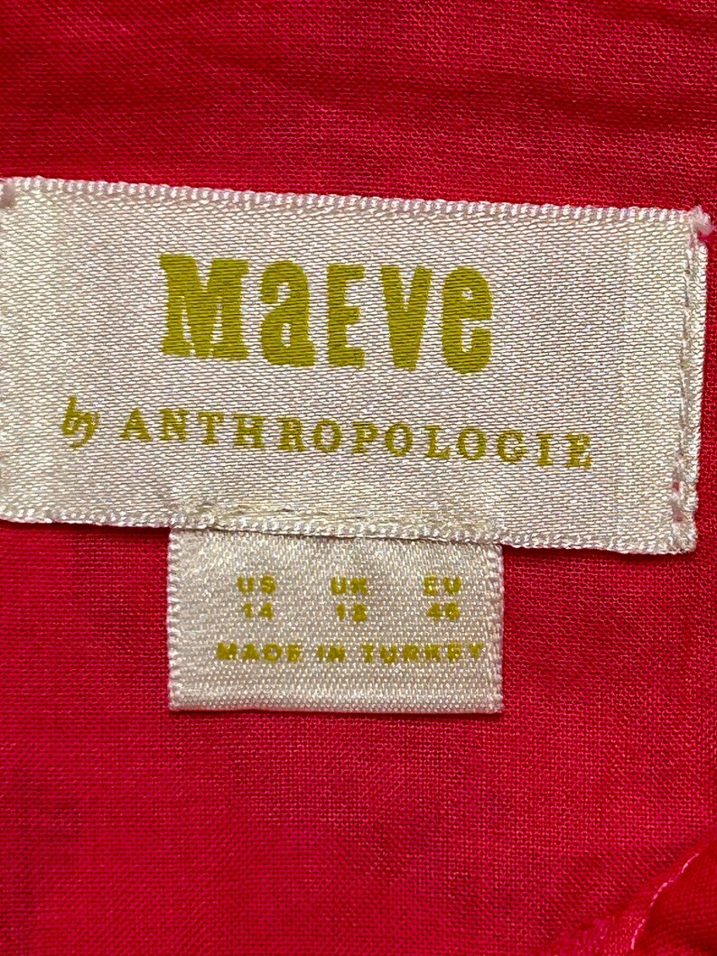 May include: White tag with gold text that reads "Maeve by Anthropologie" and size information: US 14, UK 18, EU 44. The tag is on a red fabric background.