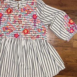 May include: A white and black striped blouse with a colourful floral embroidered design on the front and sleeves. The blouse has a round neckline and a gathered waist.