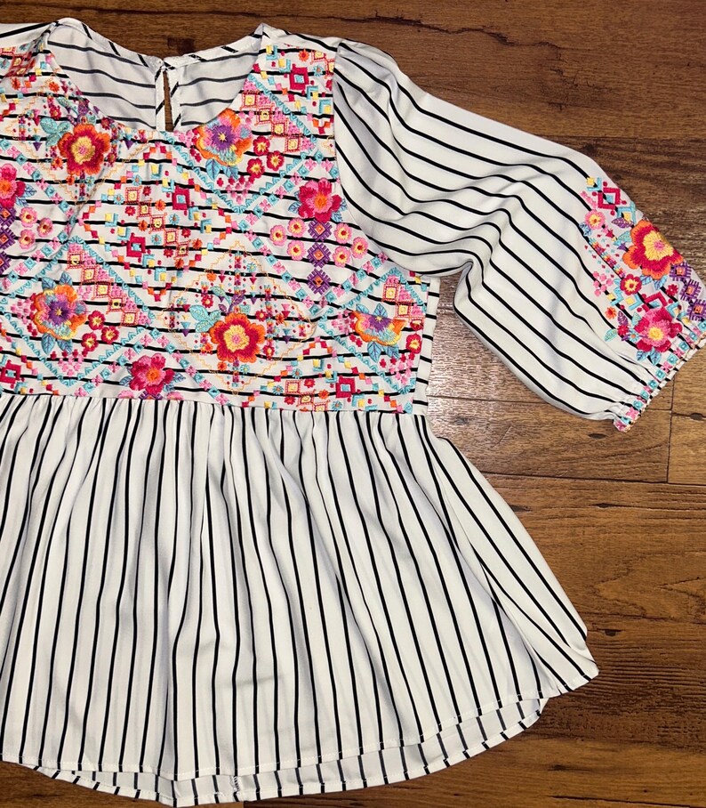 May include: A white and black striped blouse with a colourful floral embroidered design on the front and sleeves. The blouse has a round neckline and a gathered waist.