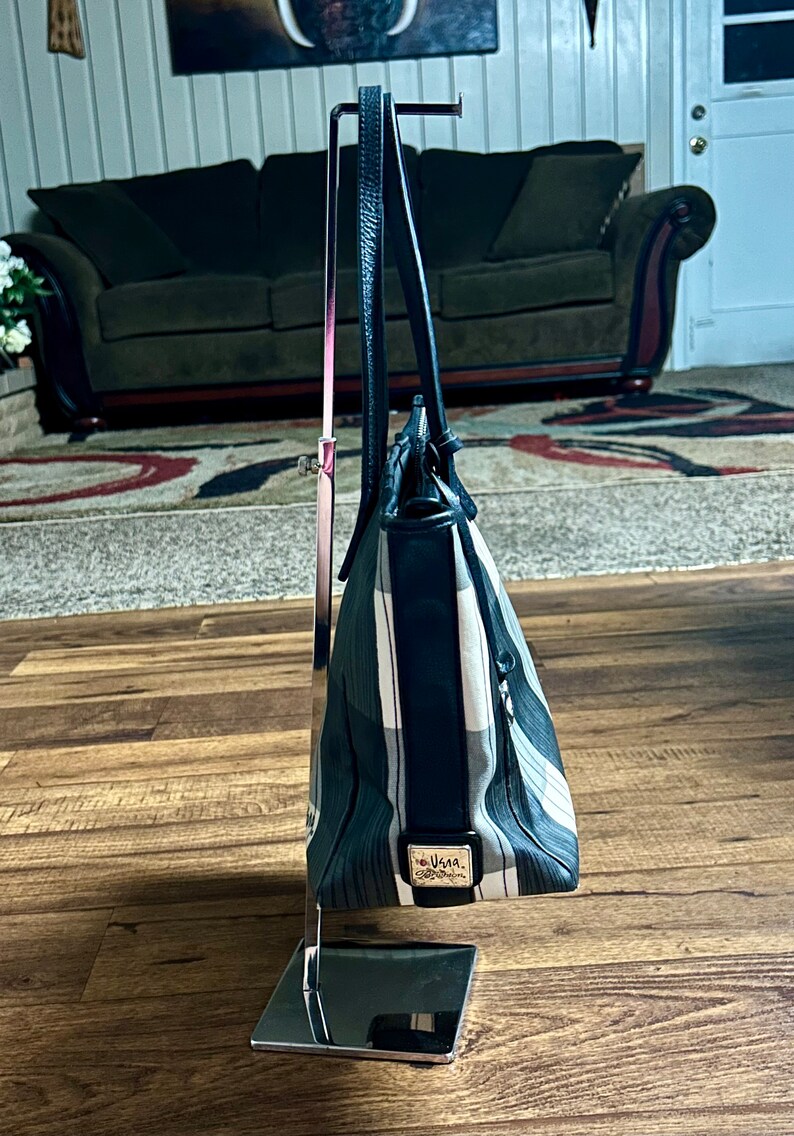 May include: A black and white striped tote bag with a black leather handle and a silver metal stand. The bag has a black leather trim and a small silver tag with the word "Vera" on it.