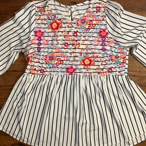 May include: White and black striped blouse with a colourful embroidered floral pattern on the front. The blouse has a ruffled hem.