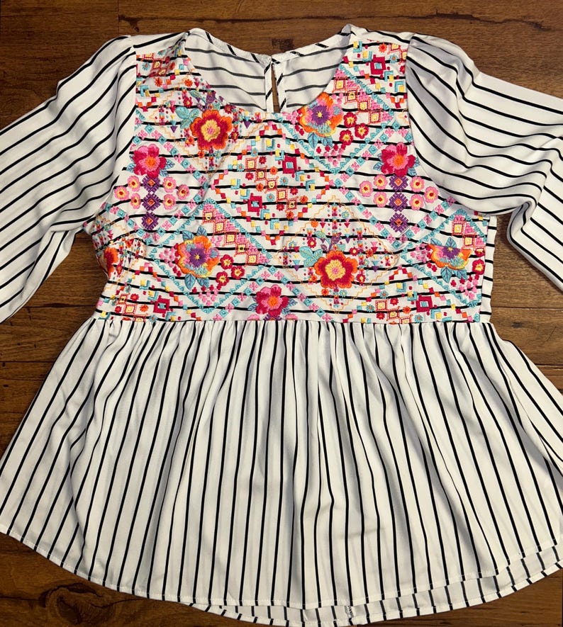 May include: White and black striped blouse with a colourful embroidered floral pattern on the front. The blouse has a ruffled hem.