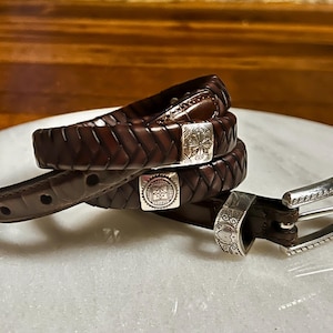 K&ouml;nnte beinhalten: Ein dunkelbrauner Lederg&uuml;rtel mit geflochtenem Design. Der G&uuml;rtel hat silberfarbene Metallschnallen mit dekorativen Mustern. Der G&uuml;rtel ist etwa 3,2 cm breit.