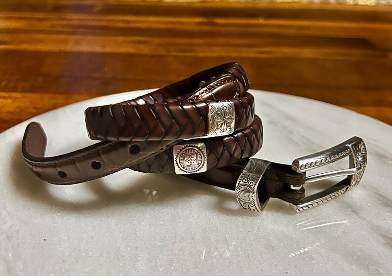 K&ouml;nnte beinhalten: Ein dunkelbrauner Lederg&uuml;rtel mit geflochtenem Design. Der G&uuml;rtel hat silberfarbene Metallschnallen mit dekorativen Mustern. Der G&uuml;rtel ist etwa 3,2 cm breit.