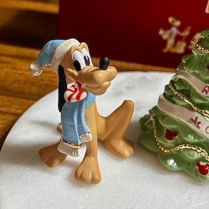 Może przedstawiać: Figurka psa Pluto w niebieskiej czapce i szaliku, trzymająca laseczkę. Po prawej stronie zielona choinka z czerwonymi ozdobami i białą wstążką. Figurka stoi na białej powierzchni.