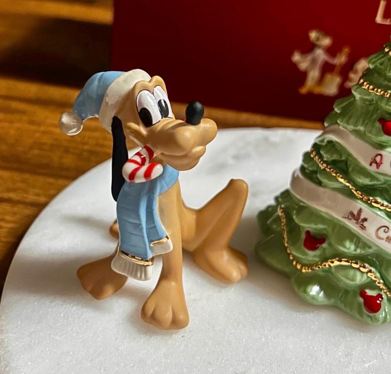 Może przedstawiać: Figurka psa Pluto w niebieskiej czapce i szaliku, trzymająca laseczkę. Po prawej stronie zielona choinka z czerwonymi ozdobami i białą wstążką. Figurka stoi na białej powierzchni.