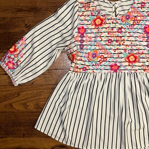 May include: White and black striped blouse with colourful embroidered floral design on the bodice.