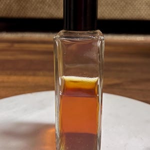 May include: A small, rectangular glass bottle with a dark brown cap. The bottle contains a layered liquid, with shades of amber and brown. The bottle is sitting on a white surface.