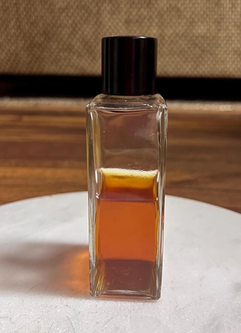 May include: A small, rectangular glass bottle with a dark brown cap. The bottle contains a layered liquid, with shades of amber and brown. The bottle is sitting on a white surface.