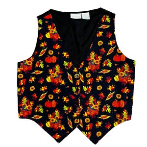 May include: A black vest featuring a vibrant autumn-themed print of pumpkins, sunflowers, corn, and colorful fall leaves. The garment has a V-neckline, a button-front closure, and a pointed hemline. A white brand label is visible at the inner neckline.
