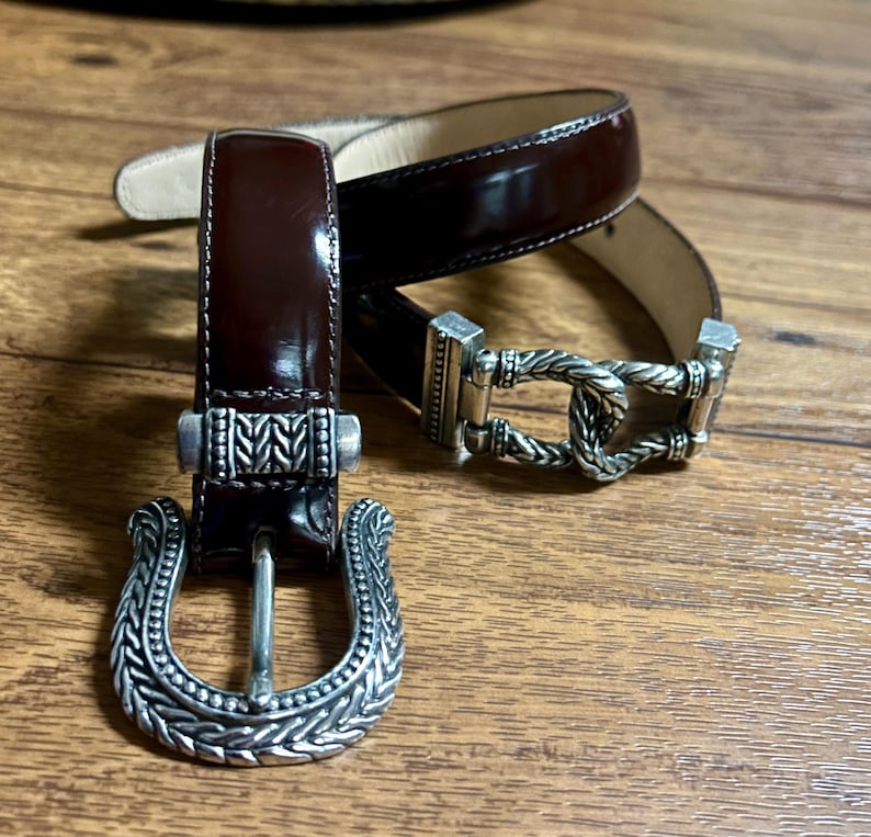 Puede incluir: Dos cinturones de cuero marr&oacute;n con hebillas plateadas. Las hebillas tienen un dise&ntilde;o intrincado con un patr&oacute;n de cuerda.