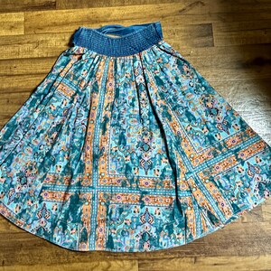 May include: A teal and orange patterned skirt with a blue elastic waistband. The skirt features a detailed, geometric design with floral and ornamental elements. The fabric has a flowing, lightweight appearance, suitable for warm weather.