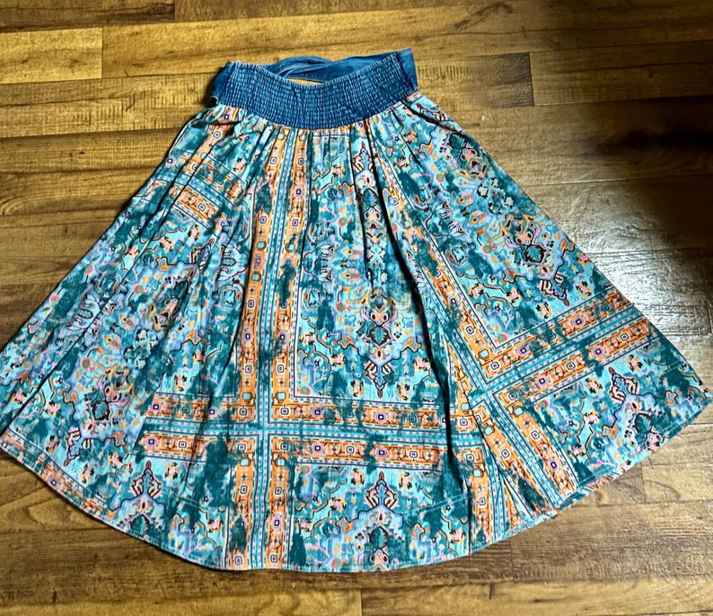 May include: A teal and orange patterned skirt with a blue elastic waistband. The skirt features a detailed, geometric design with floral and ornamental elements. The fabric has a flowing, lightweight appearance, suitable for warm weather.