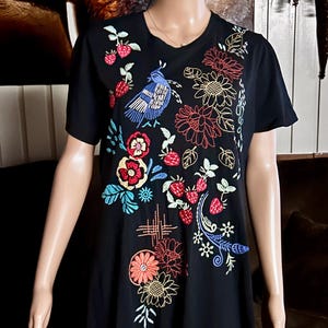 Puede incluir: Una camiseta negra de manga corta con bordados coloridos de un p&aacute;jaro azul, fresas rojas y varios patrones florales. El dise&ntilde;o cubre la parte delantera de la prenda con costuras intrincadas en tonos rojos, azules, amarillos y verdes.
