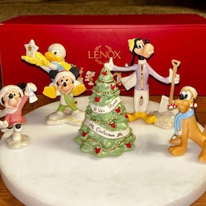 Może przedstawiać: Zestaw figurek świątecznych Lenox z postaciami Disneya. Zestaw zawiera Myszkę Miki i Minnie, Kaczora Donalda, Goofy'ego i Pluto, z udekorowaną choinką. Figurki znajdują się na białej, okrągłej podstawie, z czerwoną skrzynką Lenox w tle.