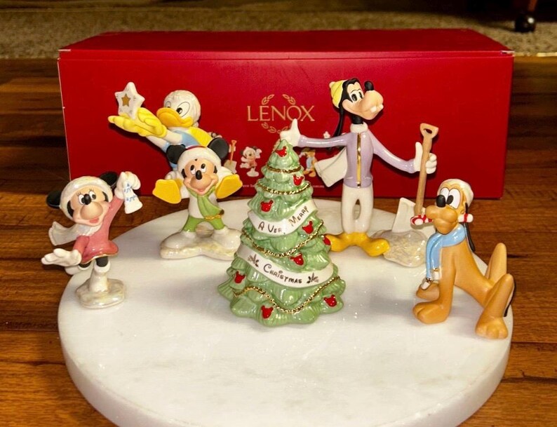 Może przedstawiać: Zestaw figurek świątecznych Lenox z postaciami Disneya. Zestaw zawiera Myszkę Miki i Minnie, Kaczora Donalda, Goofy'ego i Pluto, z udekorowaną choinką. Figurki znajdują się na białej, okrągłej podstawie, z czerwoną skrzynką Lenox w tle.