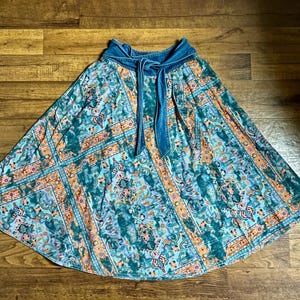 May include: A flowing skirt with a vibrant, patterned design in teal, orange, and purple hues. The skirt features a denim waistband with a tie closure. The fabric has a bohemian aesthetic, suitable for casual wear.