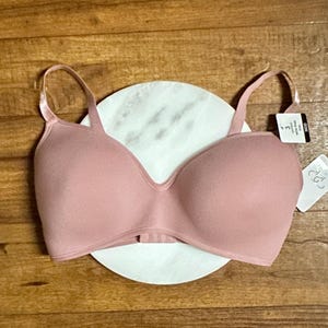 May include: A blush-colored bra with adjustable straps and a tag attached. The bra is resting on a white, circular surface with a marble pattern. The background is a wooden surface.