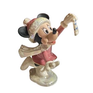 Może przedstawiać: Ceramiczna figurka Myszki Minnie w zimowym stroju. Ma na sobie czerwony płaszcz, czapkę i szalik, z czarnymi uszami i białym i niebieskim dodatkiem w dłoni. Figurka stoi na białej podstawie.