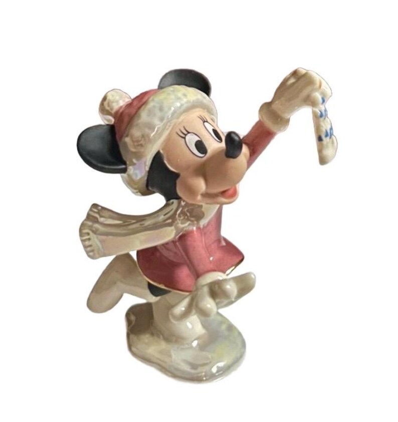 Może przedstawiać: Ceramiczna figurka Myszki Minnie w zimowym stroju. Ma na sobie czerwony płaszcz, czapkę i szalik, z czarnymi uszami i białym i niebieskim dodatkiem w dłoni. Figurka stoi na białej podstawie.