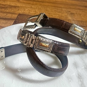 May include: Two brown leather belts with silver and gold-toned rectangular buckles. The belts have a textured, crocodile-like pattern. One belt is looped, and the other is straight, showing the buckle and belt end.