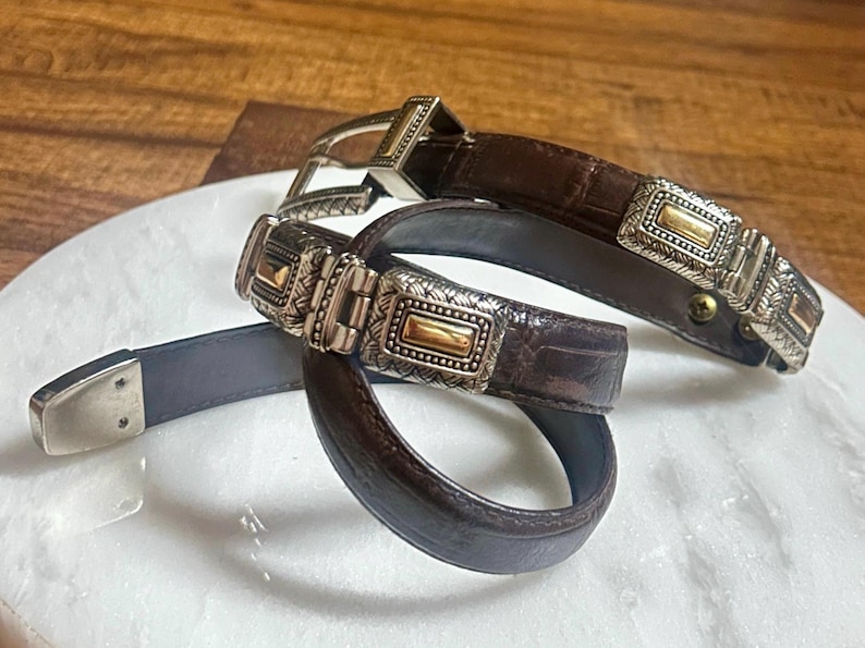 May include: Two brown leather belts with silver and gold-toned rectangular buckles. The belts have a textured, crocodile-like pattern. One belt is looped, and the other is straight, showing the buckle and belt end.