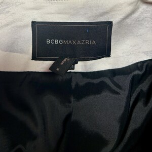 May include: Close-up of a black label with the text "BCBGMAXAZRIA" in white. A smaller tag is attached below. The interior lining is black, and the outer fabric is white with a subtle pattern. The garment appears to be a jacket or coat.