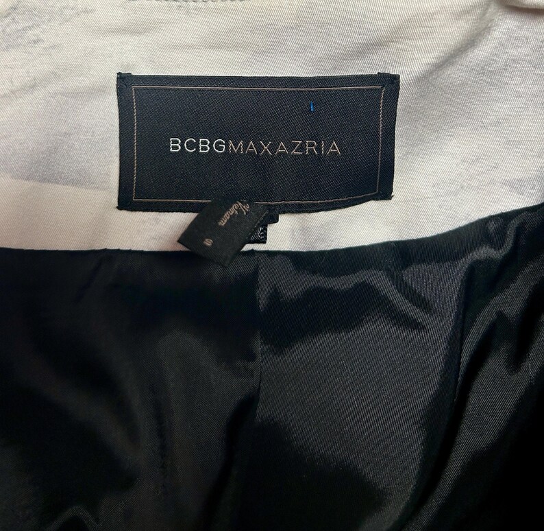 May include: Close-up of a black label with the text "BCBGMAXAZRIA" in white. A smaller tag is attached below. The interior lining is black, and the outer fabric is white with a subtle pattern. The garment appears to be a jacket or coat.