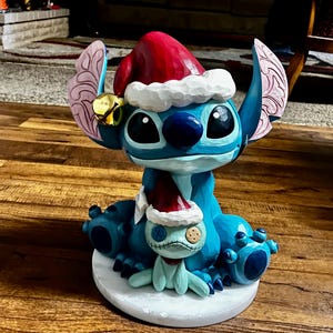 May include: A blue and white figurine of a character wearing a Santa hat, with a smaller matching figurine. The larger figurine has large black eyes and a red Santa hat with white trim. The smaller figurine is light blue with a stitched appearance and a red scarf.