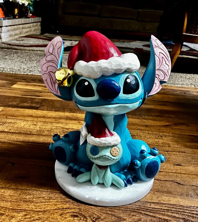 May include: A blue and white figurine of a character wearing a Santa hat, with a smaller matching figurine. The larger figurine has large black eyes and a red Santa hat with white trim. The smaller figurine is light blue with a stitched appearance and a red scarf.