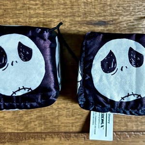 Puede incluir: Dos bolsas de dados en blanco y negro con la cara de Jack Skellington de Pesadilla antes de Navidad. Cada bolsa tiene una cara blanca con ojos negros y una boca cosida. Las bolsas est&aacute;n sobre una superficie de madera.