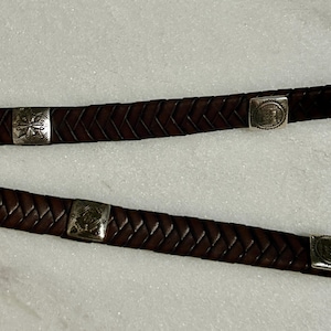 K&ouml;nnte beinhalten: Nahaufnahme von zwei braunen Lederg&uuml;rteln mit geflochtenem Design. Jeder G&uuml;rtel hat silberfarbene quadratische Schnallen mit dekorativen Mustern. Die G&uuml;rtel werden vor einem wei&szlig;en Hintergrund gezeigt.