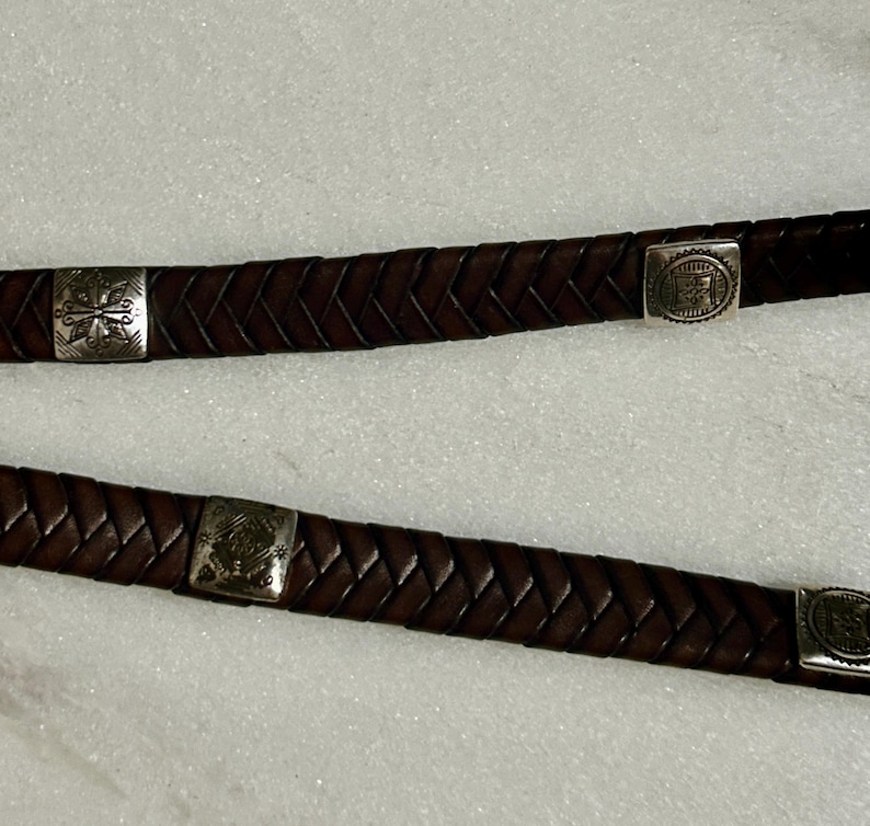 K&ouml;nnte beinhalten: Nahaufnahme von zwei braunen Lederg&uuml;rteln mit geflochtenem Design. Jeder G&uuml;rtel hat silberfarbene quadratische Schnallen mit dekorativen Mustern. Die G&uuml;rtel werden vor einem wei&szlig;en Hintergrund gezeigt.